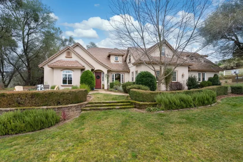 A suburban house with a landscaped front yard, featuring hedges and a pathway, set against a backdrop of trees and a partly cloudy sky.